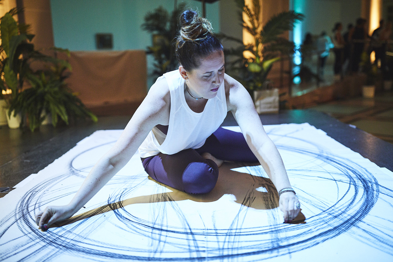 A woman practices meditative gestural drawing while sitting atop a large piece of paper, drawing large sweeping circles of charcoal around her body.
