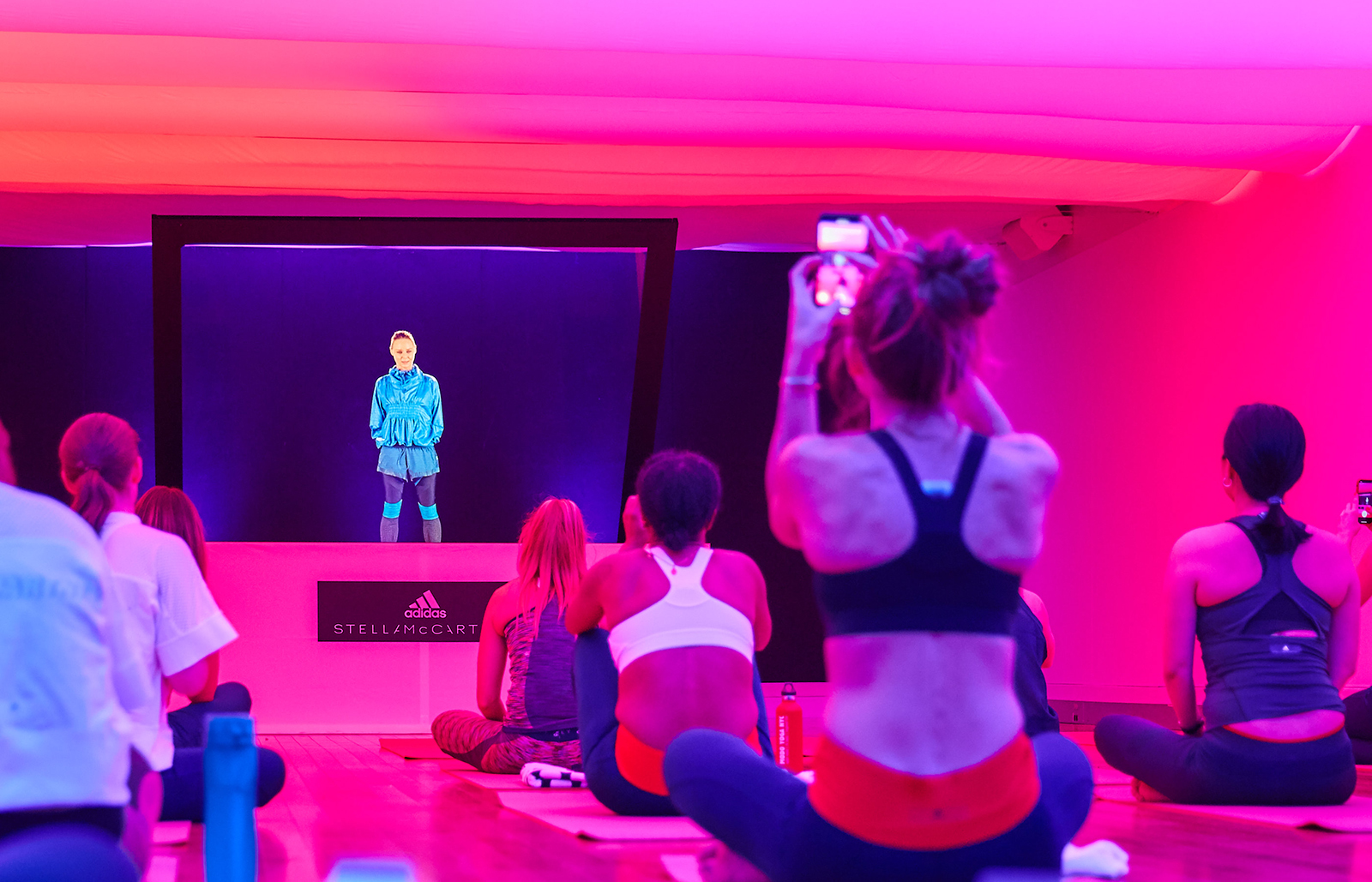 Guests to a yoga class sit cross legged on their mats in a space lit pink while they watch a hologram of an instructor.