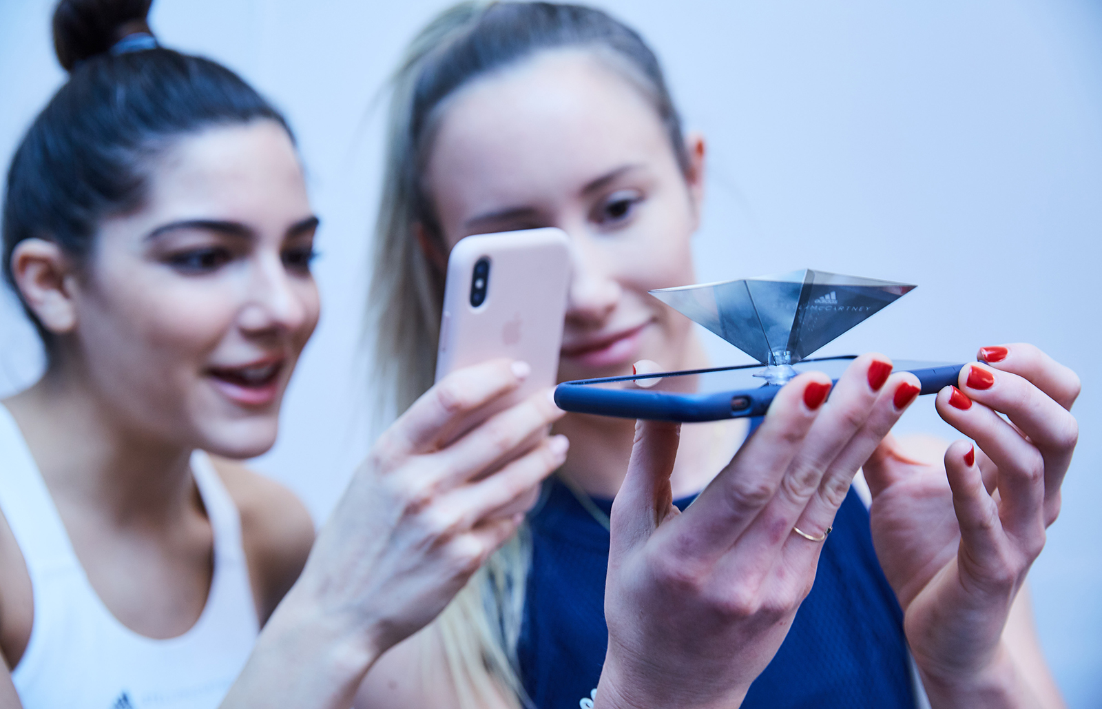 Two women look at a small mobile hologram device set up on one phone held horizontally. One woman looks at the device through the screen of a second phone.
