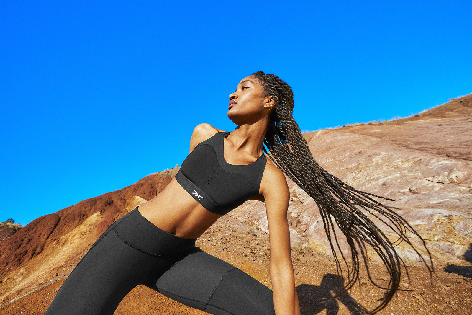 Woman wearing black bra and tights stretches in the same red earth, blue sky location.