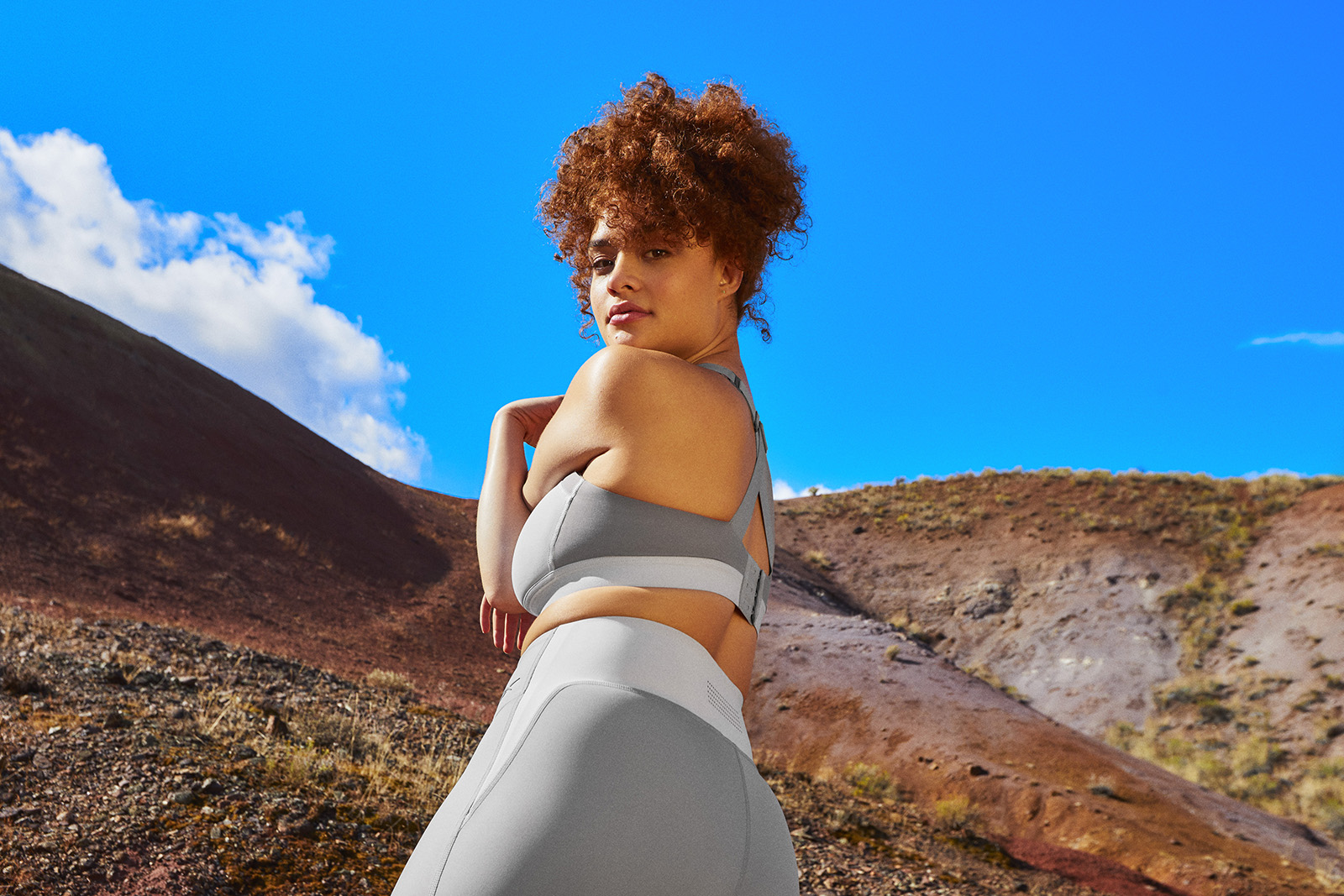 Model wearing gray bra and tights stretches her shoulder, standing on a red earth hill with brilliant blue sky behind her.