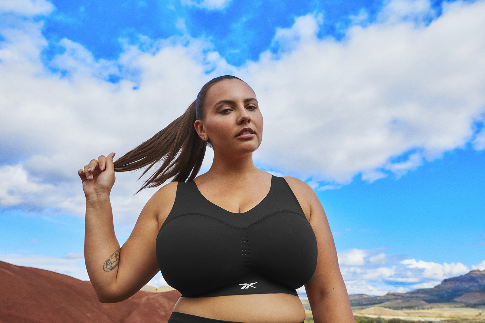 Woman wearing a black bra stands on a red hillside, with puffy clouds in a blue sky behind her.