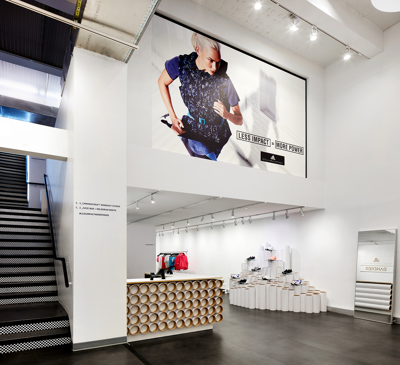 A view of the white retail space, with a cashout counter and shoe pedestals made of circular tubes of white cardboard, and a large graphic featuring Karlie Kloss that reads: Less Impact = More Power.