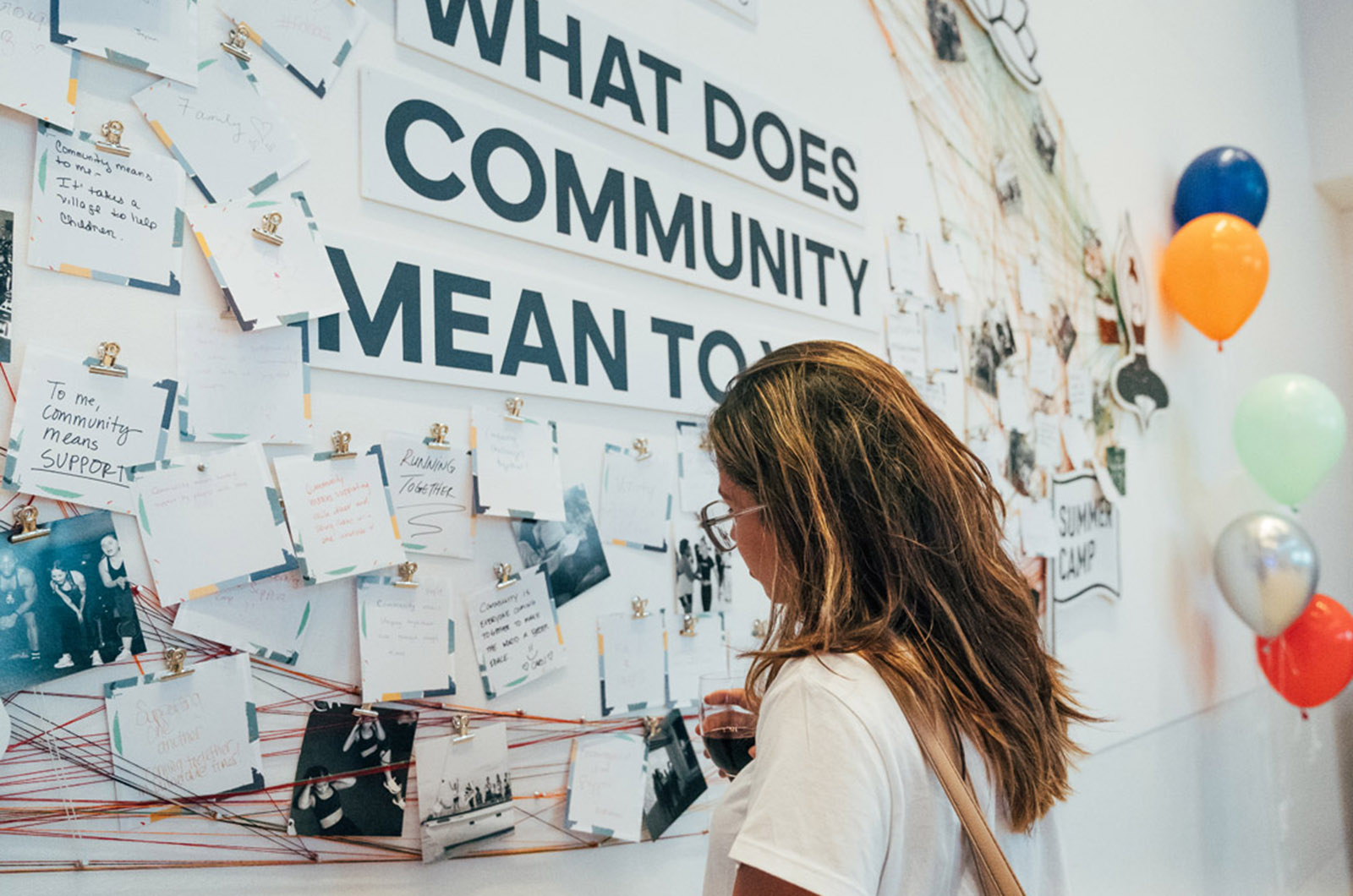 A woman looks at a wall of notes and images clipped to strings around a sign that reads: What Does Community Mean To You?