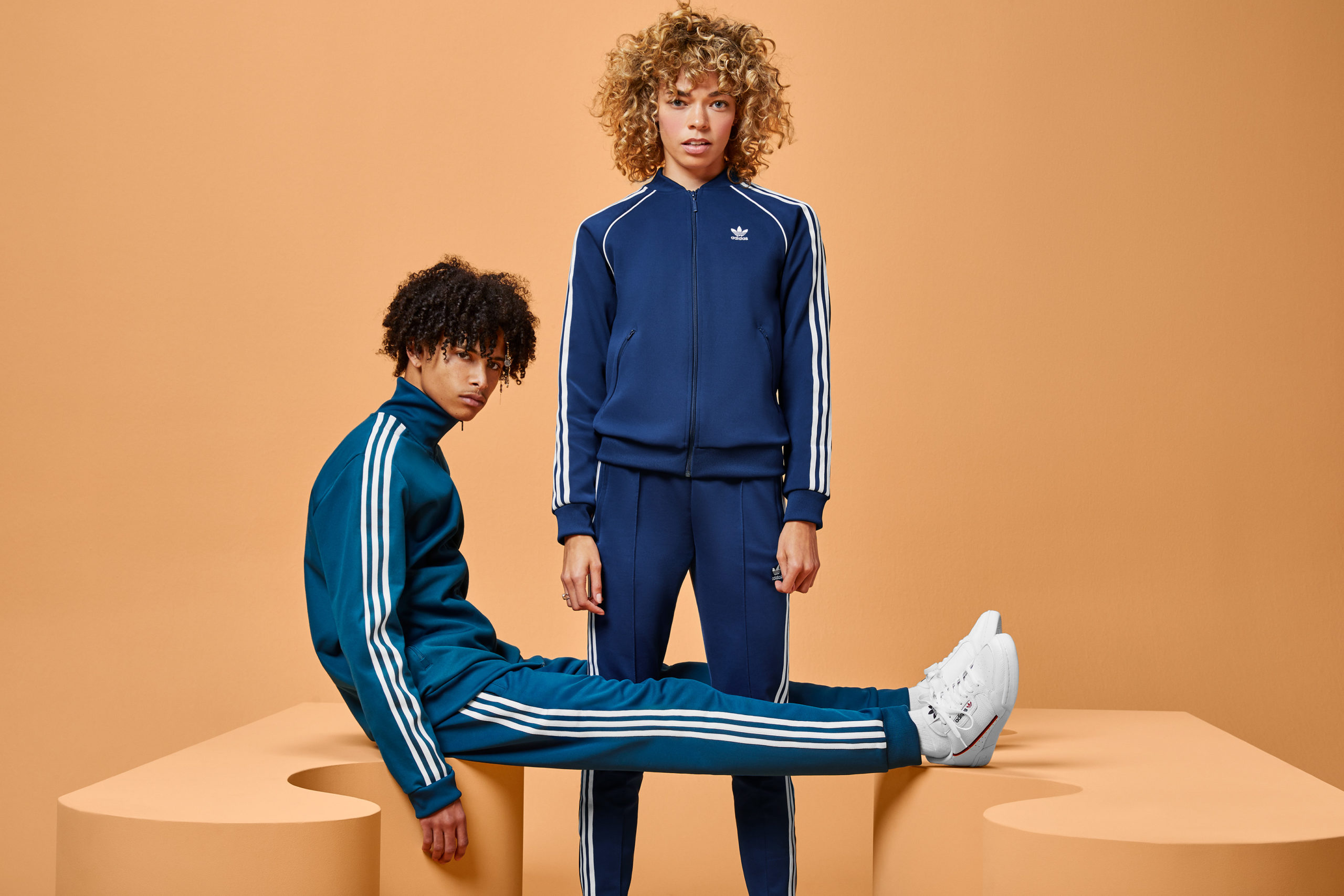 A woman wearing a blue tracksuit stands between the legs of a guy, also wearing a blue tracksuit, who’s sitting on a wavy modular shape with legs propped up on another wavy shape. Both shapes and the background are an orange-tan.