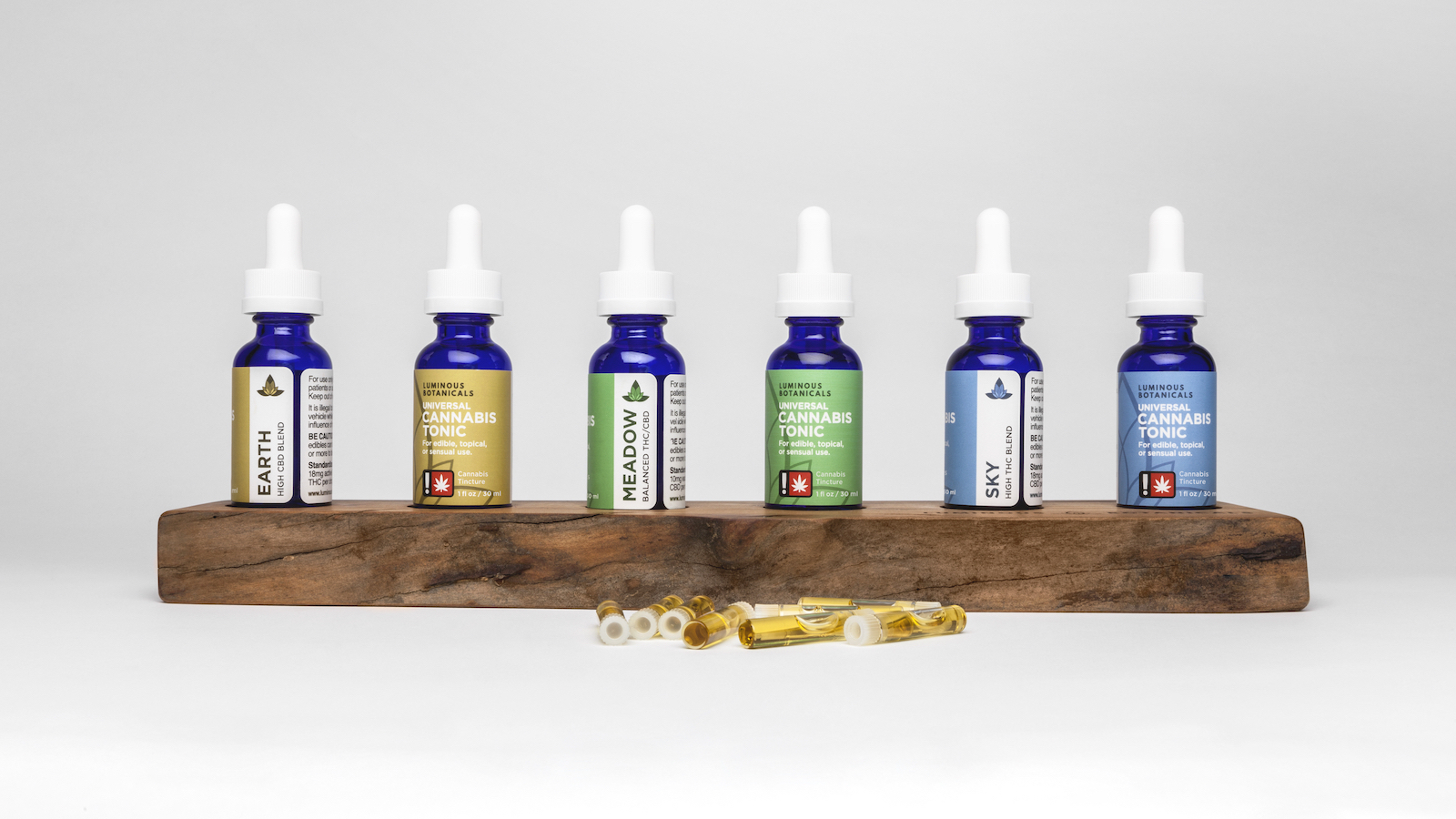 Six bottles of cannabis tonic displayed so two sides of each label can be seen. Each label has the product name listed horizontally over a color field, and the blend vertically over a white column.