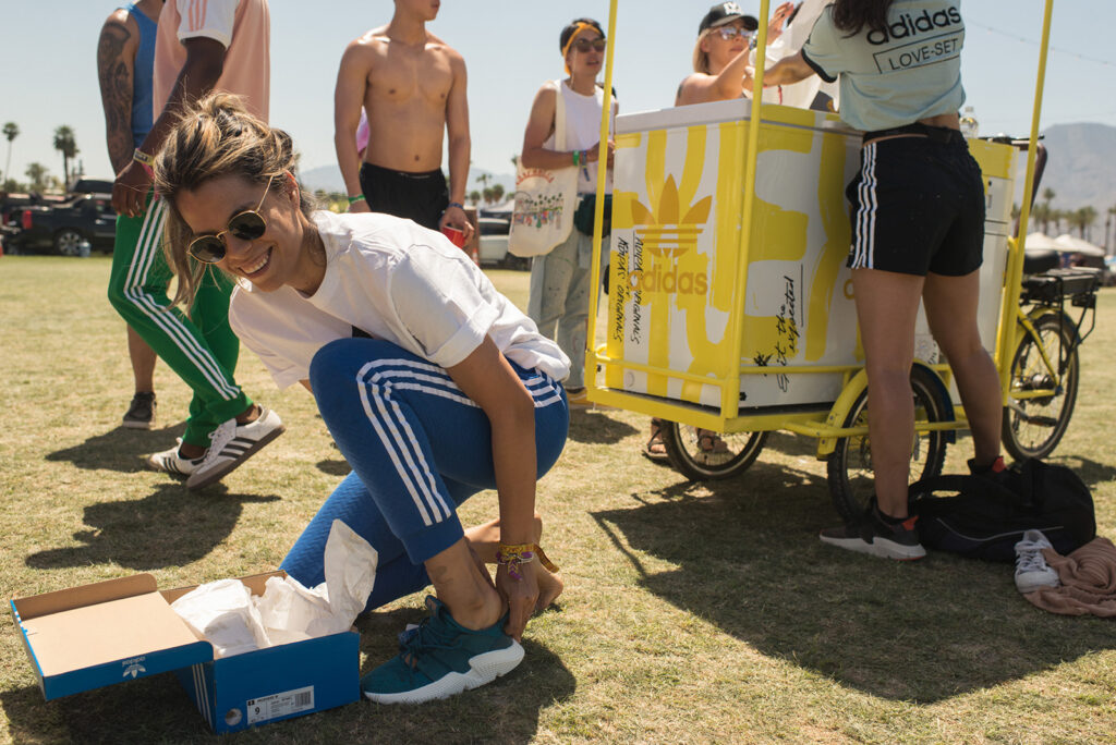 Someone kneels down to try on a sneaker, all smiles, while a few people behind them get gear being handed out from a yellow ice cream-style cart.