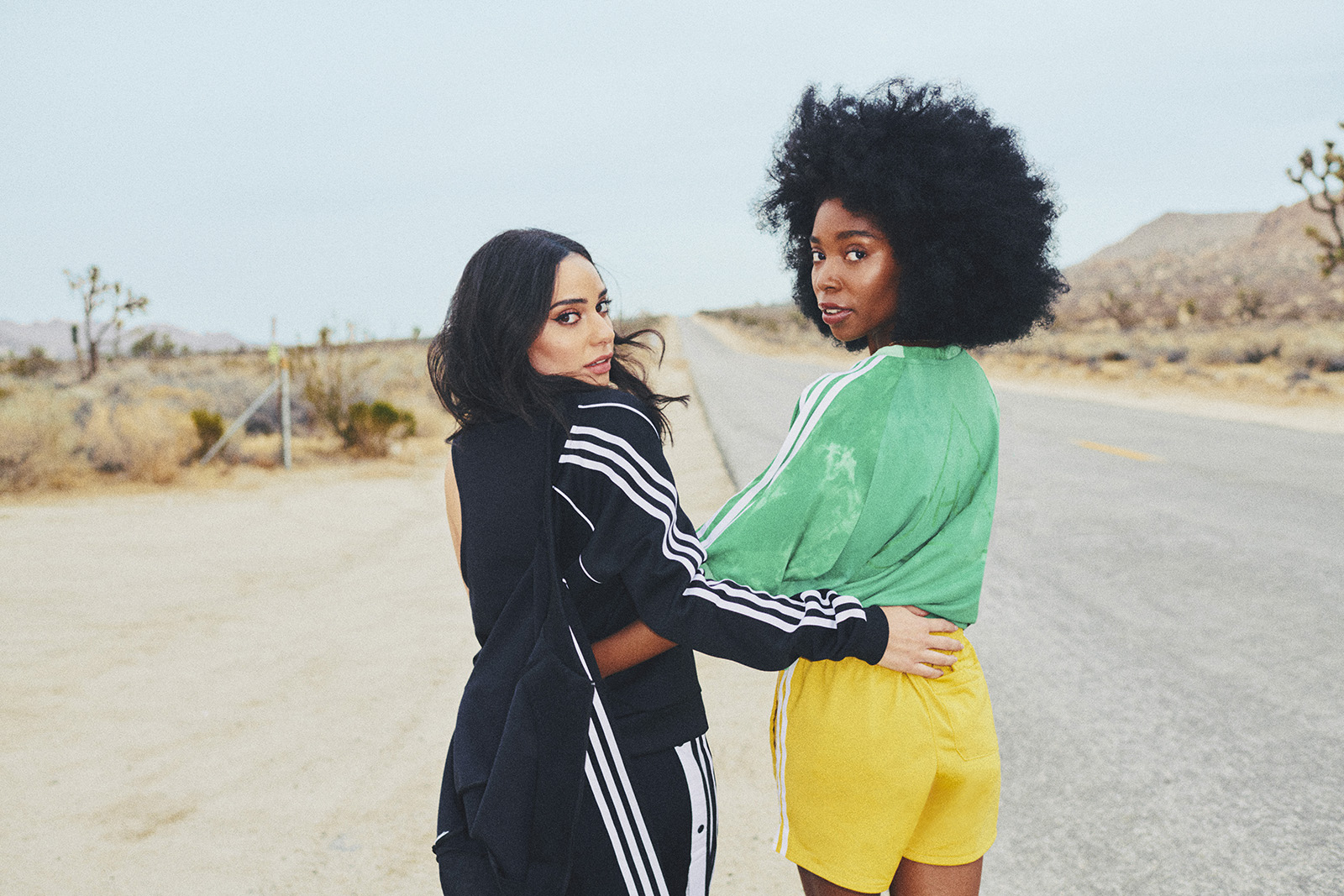 Two women, one in all black with her track jacket hanging off one shoulder and the other in bright green and yellow adidas gear stand on the side of a desert road with arms around each other’s waists, looking over their shoulders.