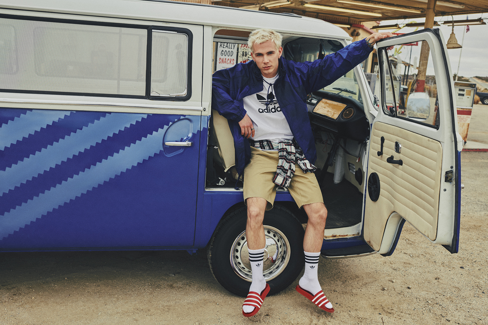 A guy wearing a blue windbreaker, adidas tee, socks, and slides leans in the open door on the passenger side of an Originals themed Volkswagen bus parked in a desert gas station.