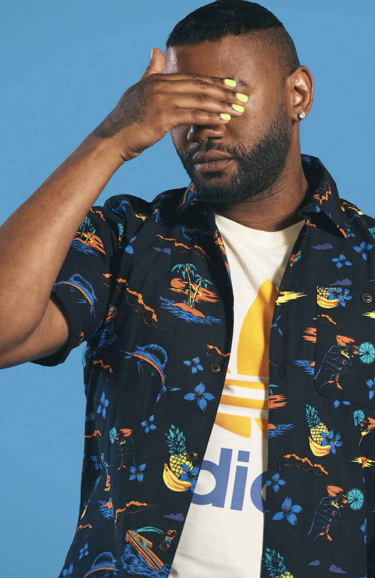 A portrait of Chanti Darling, wearing a tropical print button down tee shirt, with his hand over his eyes, nails painted bright yellow, against a blue background.
