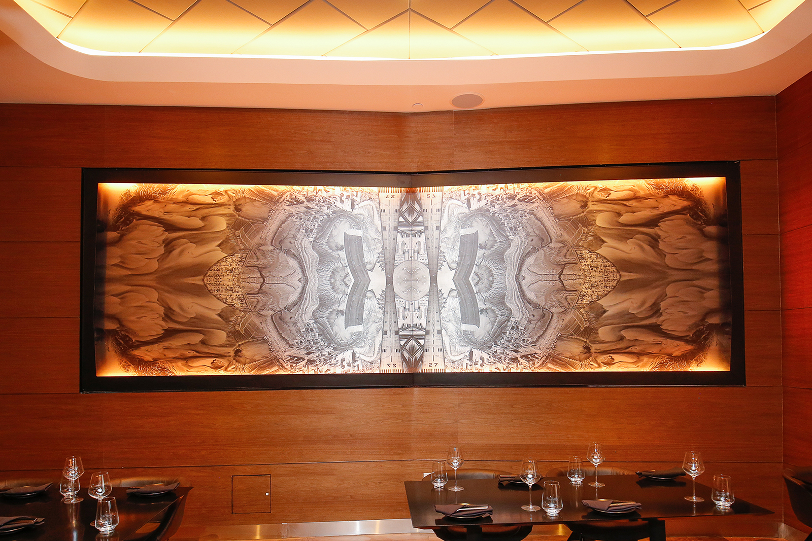 Lightbox mural of an abstracted and mirrored aerial topographical view displayed on a gently angled wood paneled wall behind set tables in the restaurant.