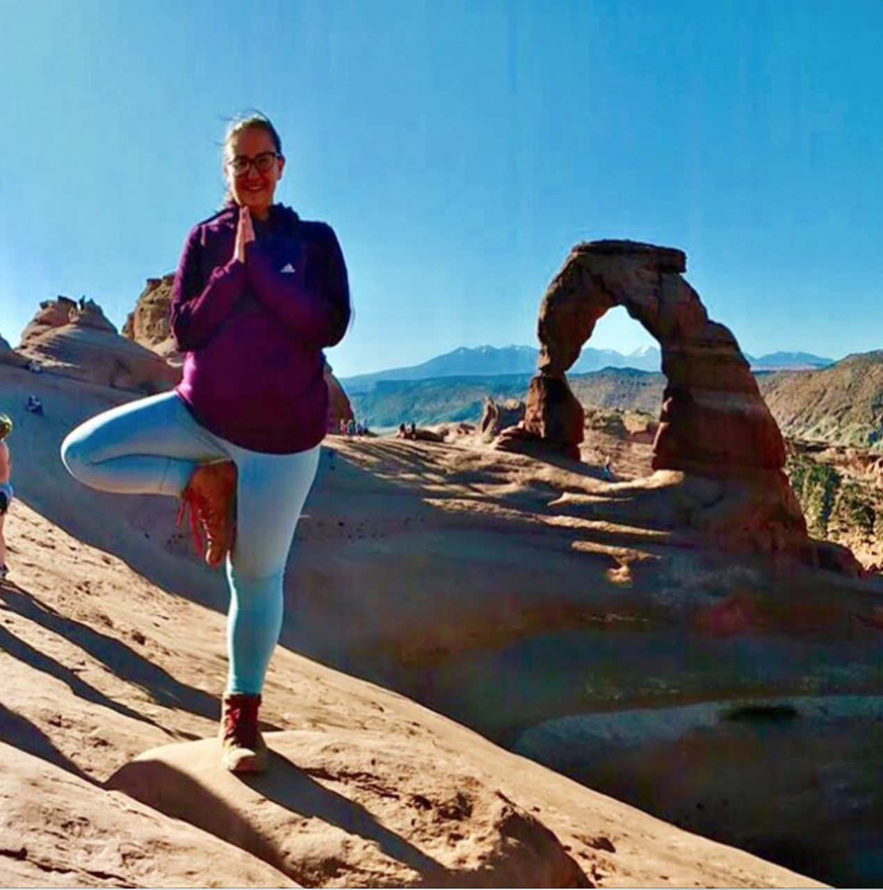 A woman does tree pose by a natural stone arch.