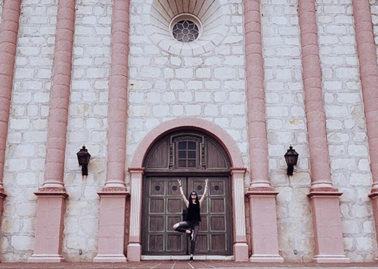 A woman does tree pose in front of the wooden door of a pink, stone building.