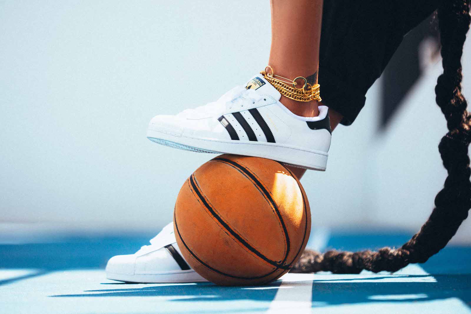 A closeup of a pair of adidas shoes on Liz Cambage, with one foot, decked out in gold anklets, resting on a basketball.