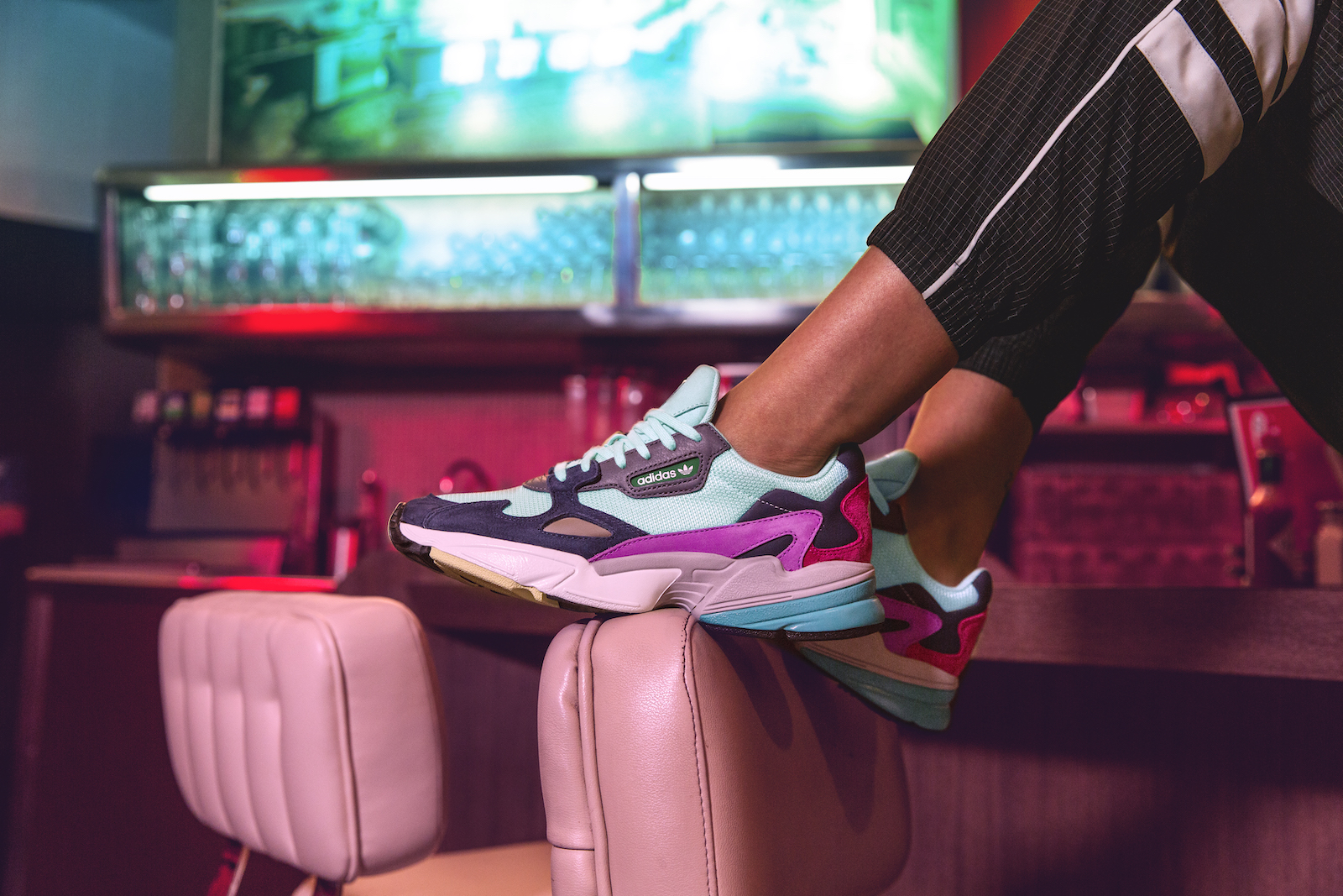 Closeup of a pair of Falcon sneakers on a model sitting on the diner counter, feet resting on the pink, leather backrest of a stool.
