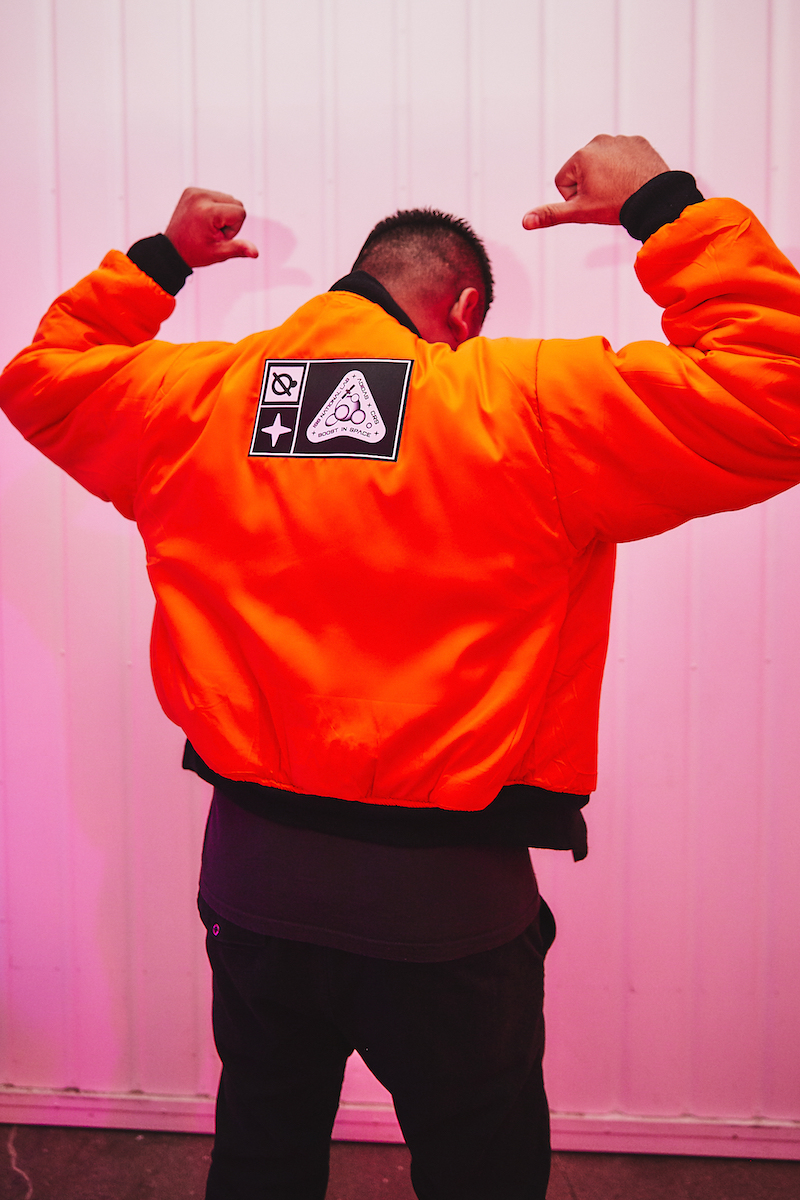 A person shows off the back of their bright orange Space Race giveaway jacket with a Boost in Space patch.