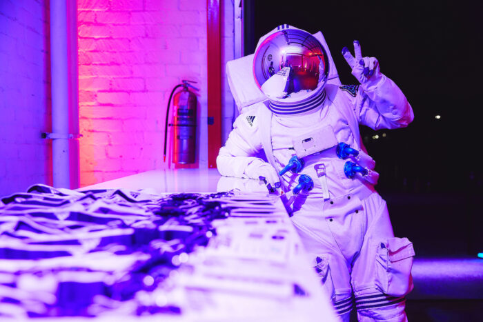 Astronaut in purple-pink light giving a peace sign while leaning on a table of event badges.