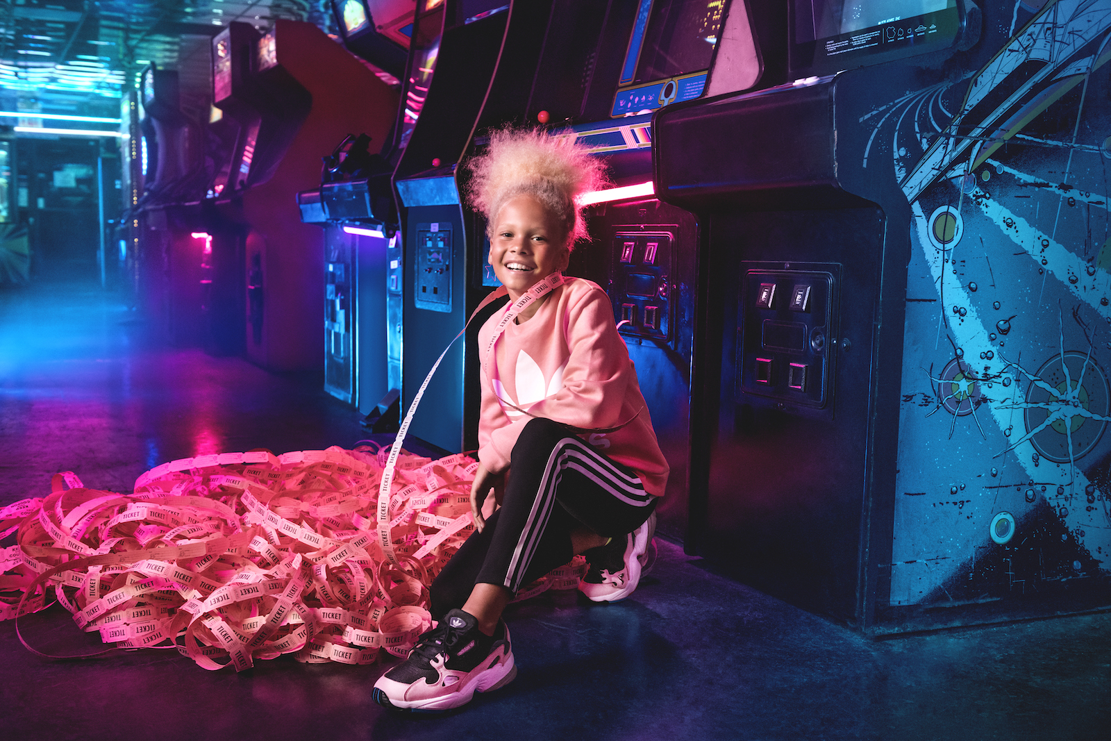 A smiling girl kneels in front of a row of arcade games with a huge pile of pink tickets.