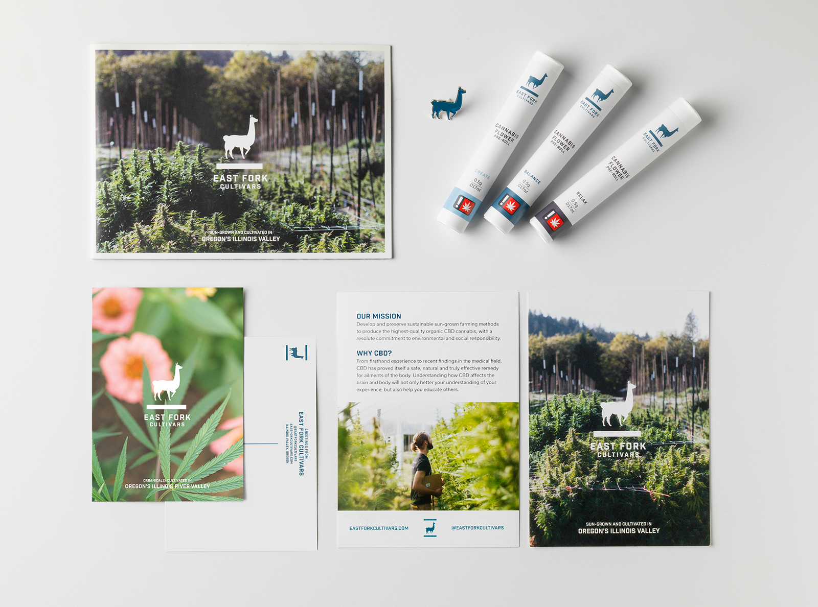 A collection of printed materials. A large card showing the plants on the farm with the white East Fork logo overtop; a blue enamel llama pin; three pre-rolls; the front and back of an informational card with a photo of the farm, stating Our Mission and Why CBD?; and the front and back of a postcard with a picture of cannabis leaves over pink flowers with the white East Fork logo overtop.