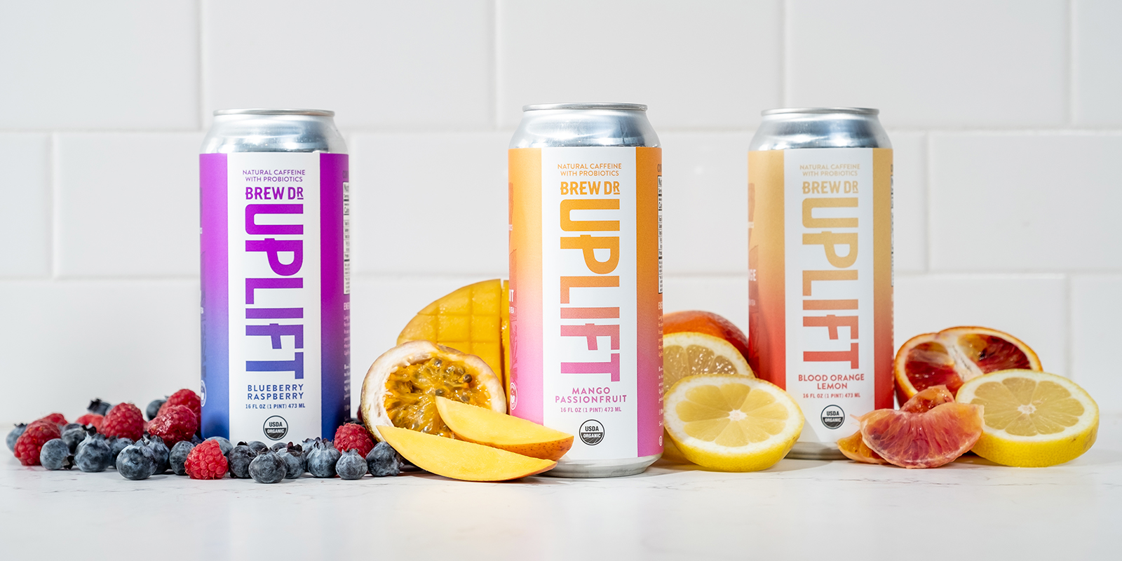 The three cans, Blueberry Raspberry, Mango Passionfruit, and Blood Orange Lemon, lined up on a counter surrounded by the fresh fruit that makes up their ingredients. Each has a white front panel with knocked-out vertical version of the Uplift logo so the gradient behind, which wraps around the whole can, shows through. The Blueberry Raspberry gradient is a dark blue to purple, the Mango Passionfruit a pink to orange, and the Blood Orange Lemon a red-orange to yellow.